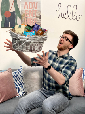 A man sits on a gray sofa in a cozy room, holding a gift basket A man sits on a gray sofa in a cozy room, holding a gift basket filled with various goodies. He is wearing a plaid shirt and glasses, with a cheerful expression as he seems to be playfully tossing the basket in the air. The room is decorated with pillows