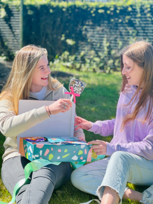In a sunlit garden, two young women are seated on the grass, joyfully exploring a colorful gift box. In a sunlit garden, two young women are seated on the grass, joyfully exploring a colorful gift box. The woman on the left, dressed in a beige hoodie, is laughing as she holds up a heart-shaped chocolate with a pink ribbon. The woman on the right, in a la